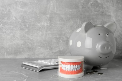 Educational dental typodont model, piggy bank, coins and calculator on grey table, space for text. Expensive treatment