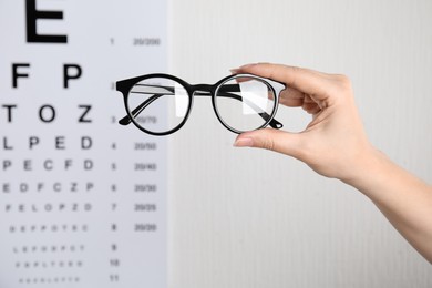 Woman holding glasses against eye chart on light background, closeup. Ophthalmologist prescription