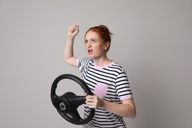 Emotional young woman with steering wheel on grey background