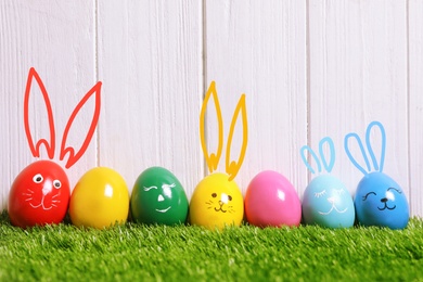 Several eggs with drawn faces and ears as Easter bunnies among others on green grass against white wooden background