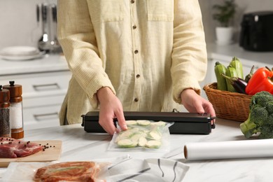Woman using sealer for vacuum packing with plastic bag of zucchini at white table in kitchen, closeup