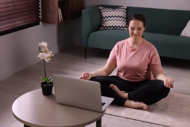 Overweight woman meditating on rug while watching online class at home