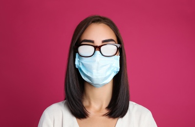 Young woman with foggy glasses caused by wearing disposable mask on pink background. Protective measure during coronavirus pandemic