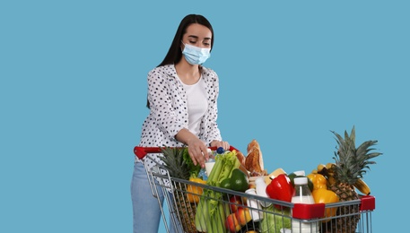 Young woman in medical mask with shopping cart full of groceries on light blue background
