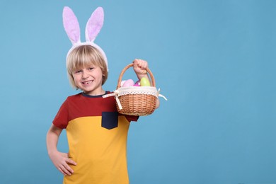 Photo of Happy boy in bunny ears headband holding wicker basket with painted Easter eggs on turquoise background. Space for text