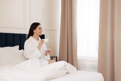 Happy young woman wearing bathrobe drinking coffee on bed in hotel room