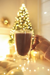 Woman with cup of drink and blurred Christmas tree on background, closeup