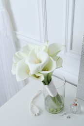 Photo of Beautiful calla lily flowers tied with ribbon in glass vase, bottle of perfume and jewelry on white table