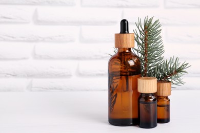 Bottles of essential oil and pine branch on white wooden table near brick wall, space for text