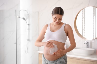 Young pregnant woman applying cosmetic product on belly in bathroom