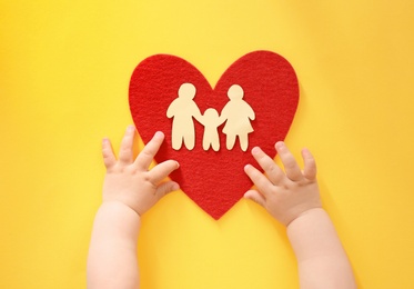 Little child's hands near red heart and paper silhouette of people on color background, top view