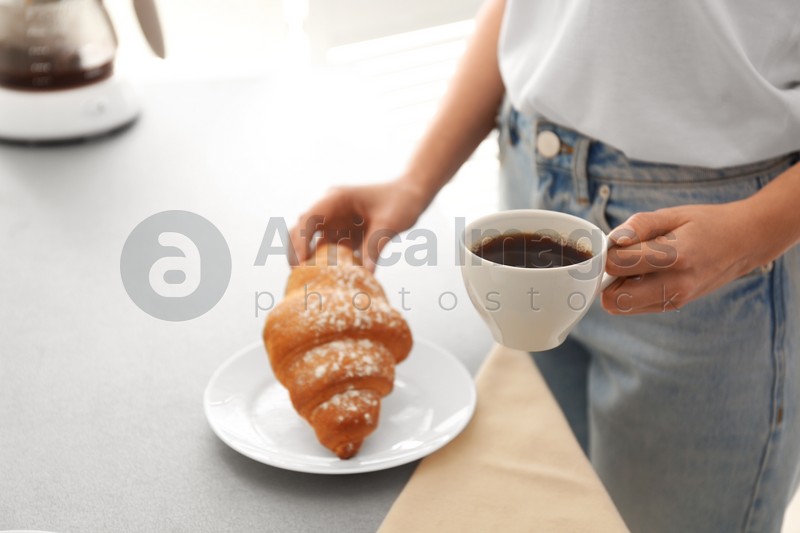 Woman having tasty breakfast with croissant and coffee at home, closeup. Morning routine