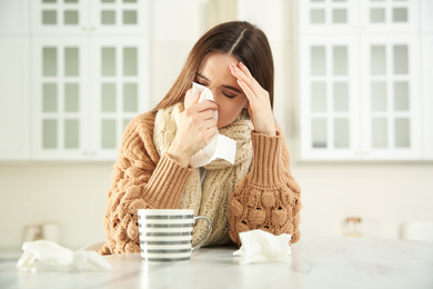 Sick young woman with cup of hot drink and tissues in kitchen. Influenza virus
