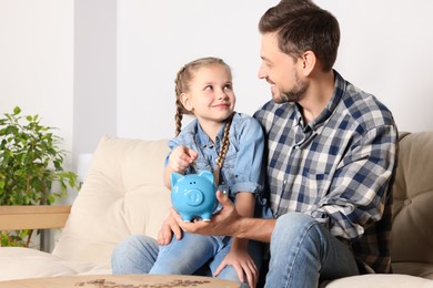 Photo of Little girl with her father putting coin into piggy bank at home