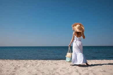 Woman with beach bag and straw hat on sand near sea, back view