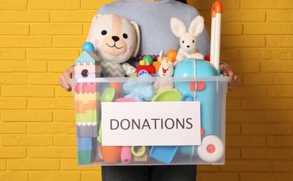 Woman holding donation box full of different toys near yellow brick wall, closeup