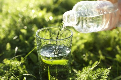 Woman pouring fresh water from bottle into glass on green grass outdoors, closeup