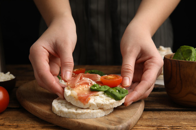 Woman holding puffed rice cake with prosciutto, tomato and basil at wooden table, closeup