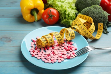 Plate with weight loss pills, measuring tape and fork near pile of vegetables on light blue wooden table