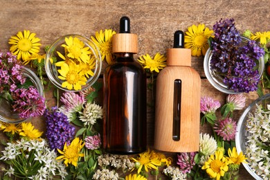 Bottles of essential oils surrounded by beautiful flowers on wooden table, flat lay