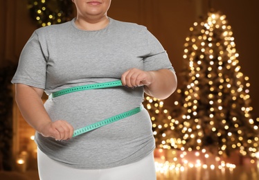 Overweight woman measuring her waist in room with Christmas tree after holidays, closeup
