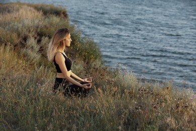 Young woman meditating near river on sunny day, space for text