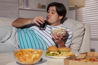 Overweight man with bowl of popcorn and drink on sofa at home