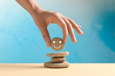Woman putting stone with drawn happy face on stack against light blue background, closeup. Zen concept