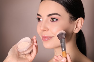 Professional makeup artist applying powder onto beautiful young woman's face with brush on dusty rose background, closeup