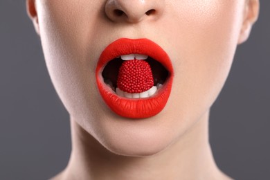Young woman with beautiful red lips makeup eating candy on grey background, closeup