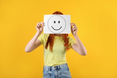 Photo of Woman hiding behind sheet of paper with happy face on yellow background