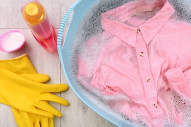 Pink shirt in basin, powder, gloves and bottle of detergent on floor, flat lay. Hand washing laundry