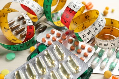 Many different weight loss pills and measuring tape on white wooden table, closeup