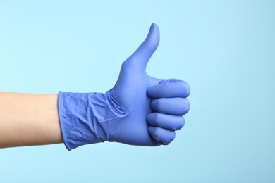 Person in medical gloves showing thumb up on light blue background, closeup of hand