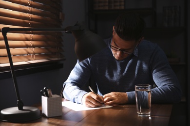 Man writing letter at wooden table in dark room
