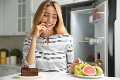 Woman choosing between cake and healthy fruits at table in kitchen