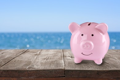 Saving money for summer vacation. Piggy bank on wooden surface near sea, space for text