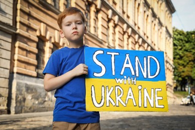 Sad boy holding poster in colors of national flag and words Stand with Ukraine on city street