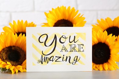 Card with life-affirming phrase You Are Amazing and sunflowers on light table against white brick wall