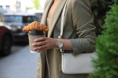 Photo of Woman holding tasty croissant and cup of coffee outdoors, closeup
