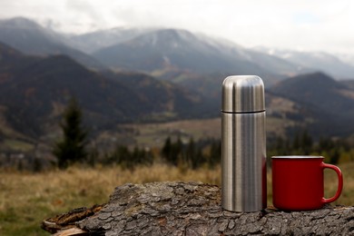 Silver thermos and mug on log in mountains, space for text