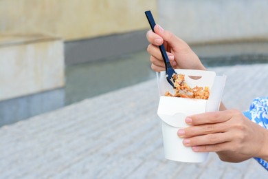 Photo of Woman eating takeaway noodles from paper box with fork outdoors, closeup and space for text. Street food