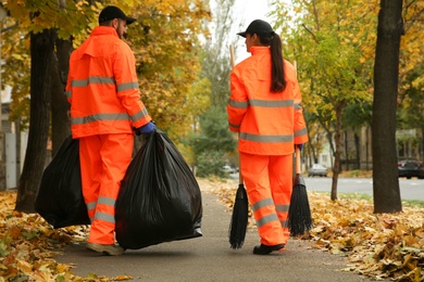 Street cleaners with brooms and garbage bags outdoors on autumn day, back view