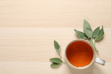 Cup of aromatic sage tea and fresh leaves on wooden table, flat lay. Space for text