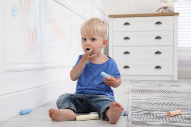 Photo of Mischievous little boy near white wall with colorful chalk scribbles at home