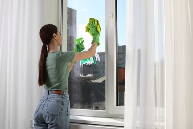 Young woman cleaning window glass with rag and detergent at home, space for text