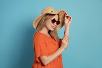 Beautiful young woman wearing straw hat and sunglasses on light blue background. Stylish headdress