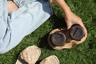 Woman holding takeaway cardboard coffee cups with plastic lids on green grass, top view