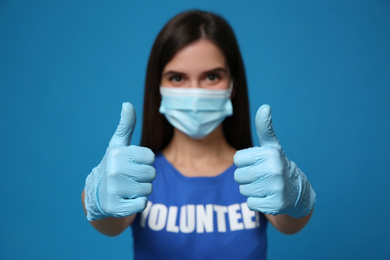 Female volunteer in protective mask and gloves showing thumbs up gesture against blue background, focus on hands. Aid during coronavirus quarantine	