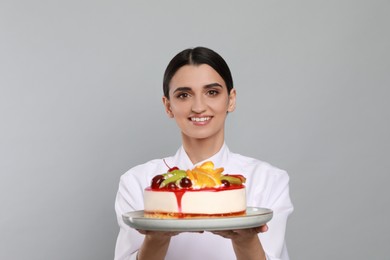 Photo of Happy professional confectioner in uniform holding delicious cake on light grey background